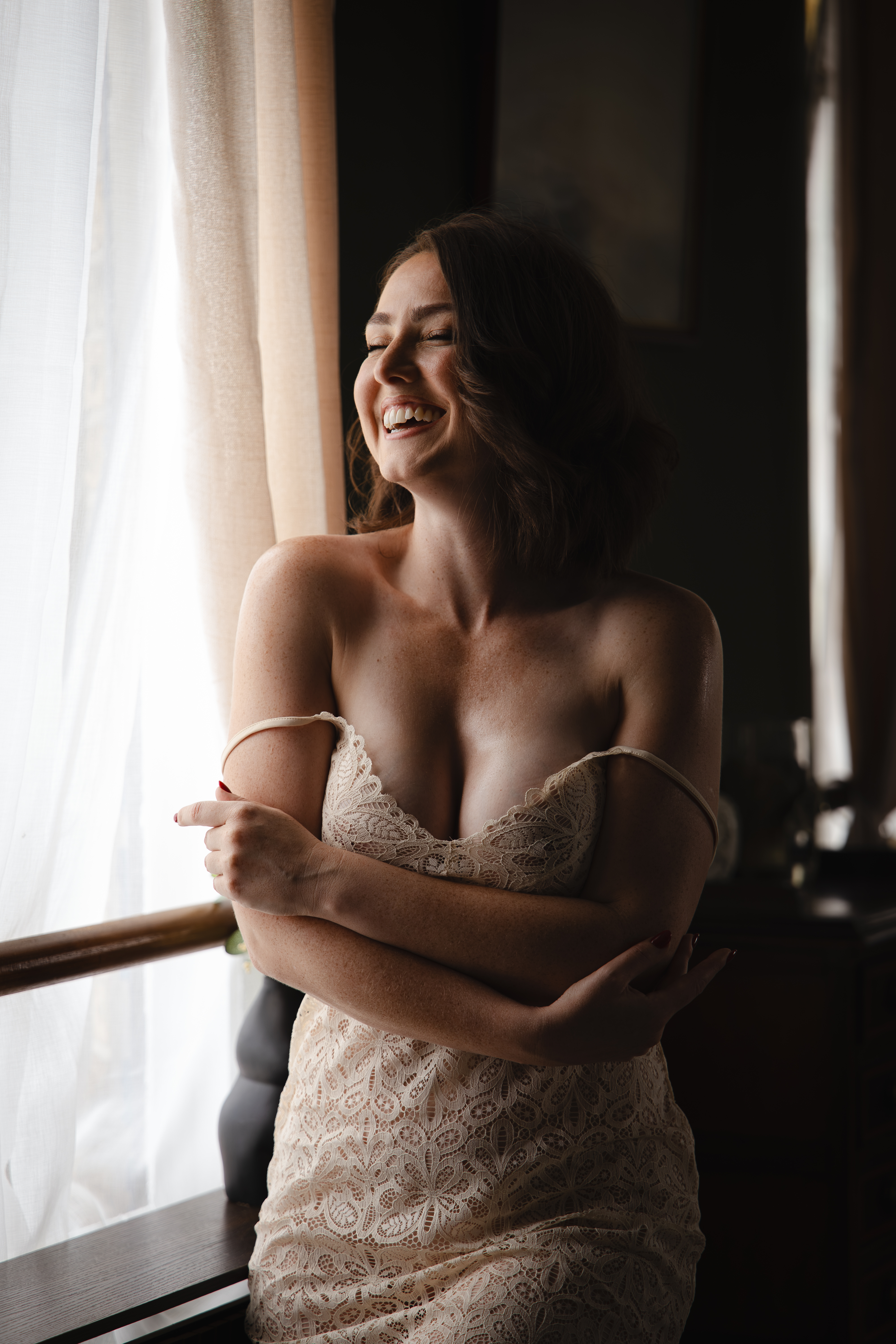 A woman laughing beside the studio window in a cream lace outfit